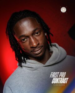 Portrait of a man with dreadlocks wearing a gray hoodie against a red backdrop; 'FIRST PRO CONTRACT' logo and 'Elisha Boateng' text visible.