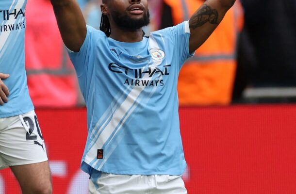 Manchester City player celebrates on the pitch with arms raised, crowd in background.