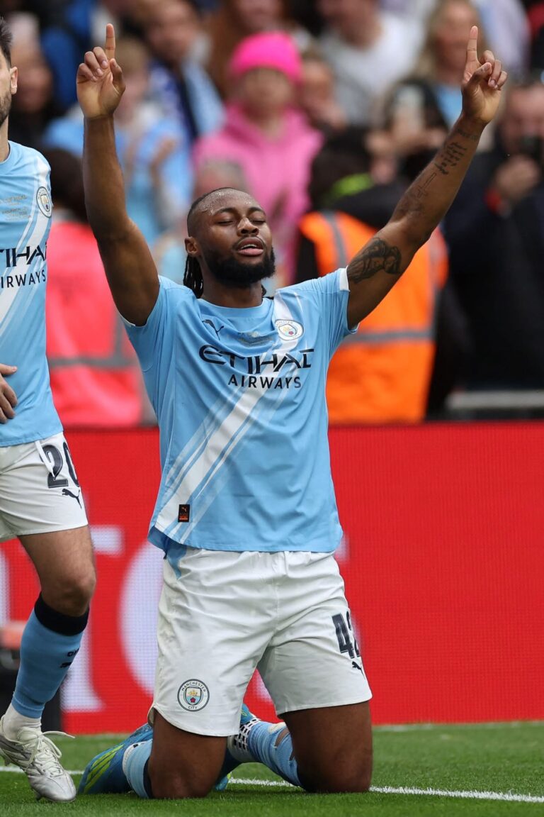 Manchester City player celebrates on the pitch with arms raised, crowd in background.