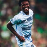 Soccer player in a light blue jersey celebrating with a big smile on the field during a match.