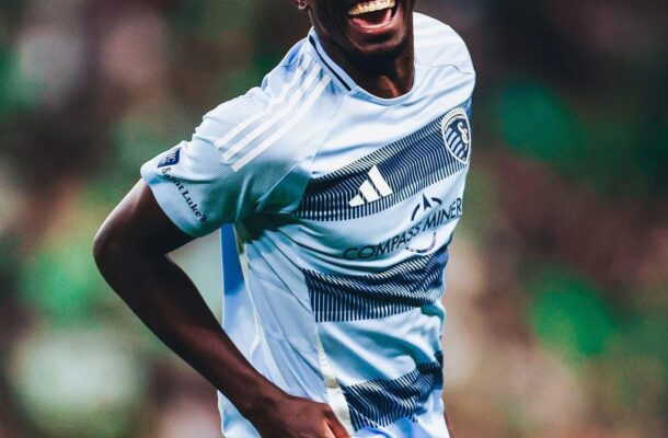 Soccer player in a light blue jersey celebrating with a big smile on the field during a match.