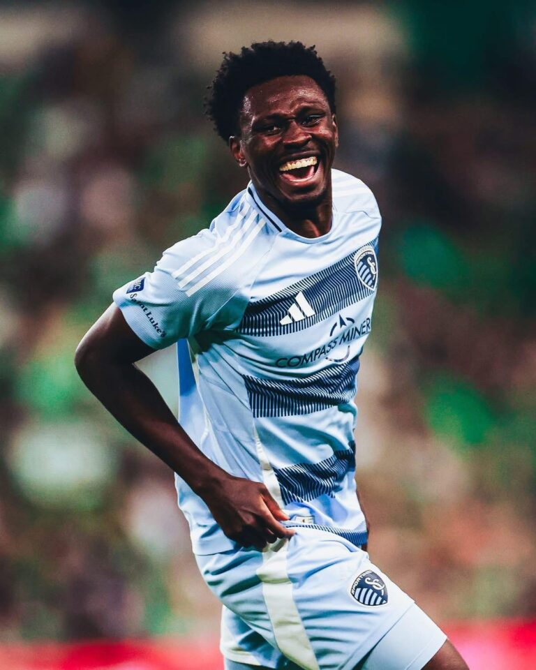 Soccer player in a light blue jersey celebrating with a big smile on the field during a match.