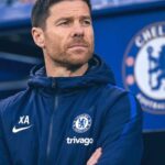 Chelsea FC staff member in a navy training jacket with Crest and Trivago logo, standing with arms crossed and a thoughtful expression.