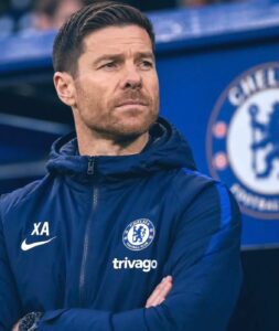Chelsea FC staff member in a navy training jacket with Crest and Trivago logo, standing with arms crossed and a thoughtful expression.