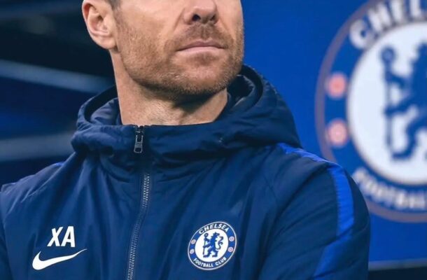 Chelsea FC staff member in a navy training jacket with Crest and Trivago logo, standing with arms crossed and a thoughtful expression.