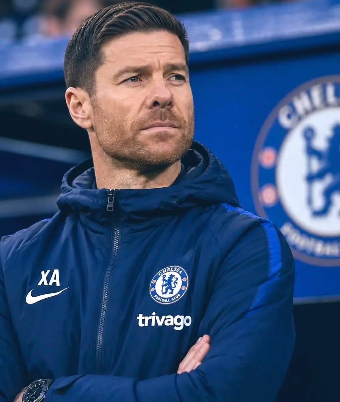 Chelsea FC staff member in a navy training jacket with Crest and Trivago logo, standing with arms crossed and a thoughtful expression.