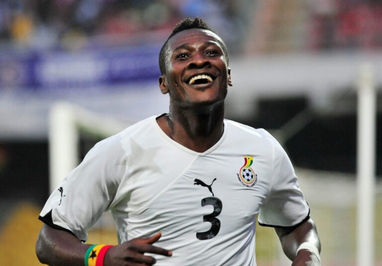 Football player in a white jersey celebrating, smiling during a match with Senegal crest visible on chest and a rainbow wristband.