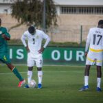 Soccer players on a grassy field: a player in white jersey number 3 stands over a ball, flanked by players in green uniforms during a match.