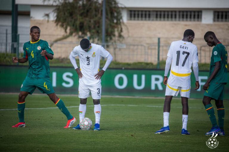 Soccer players on a grassy field: a player in white jersey number 3 stands over a ball, flanked by players in green uniforms during a match.