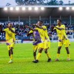 Soccer players in bright yellow jerseys celebrate on the field with a cheering crowd in the stands behind them.