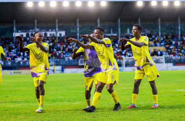 Soccer players in bright yellow jerseys celebrate on the field with a cheering crowd in the stands behind them.