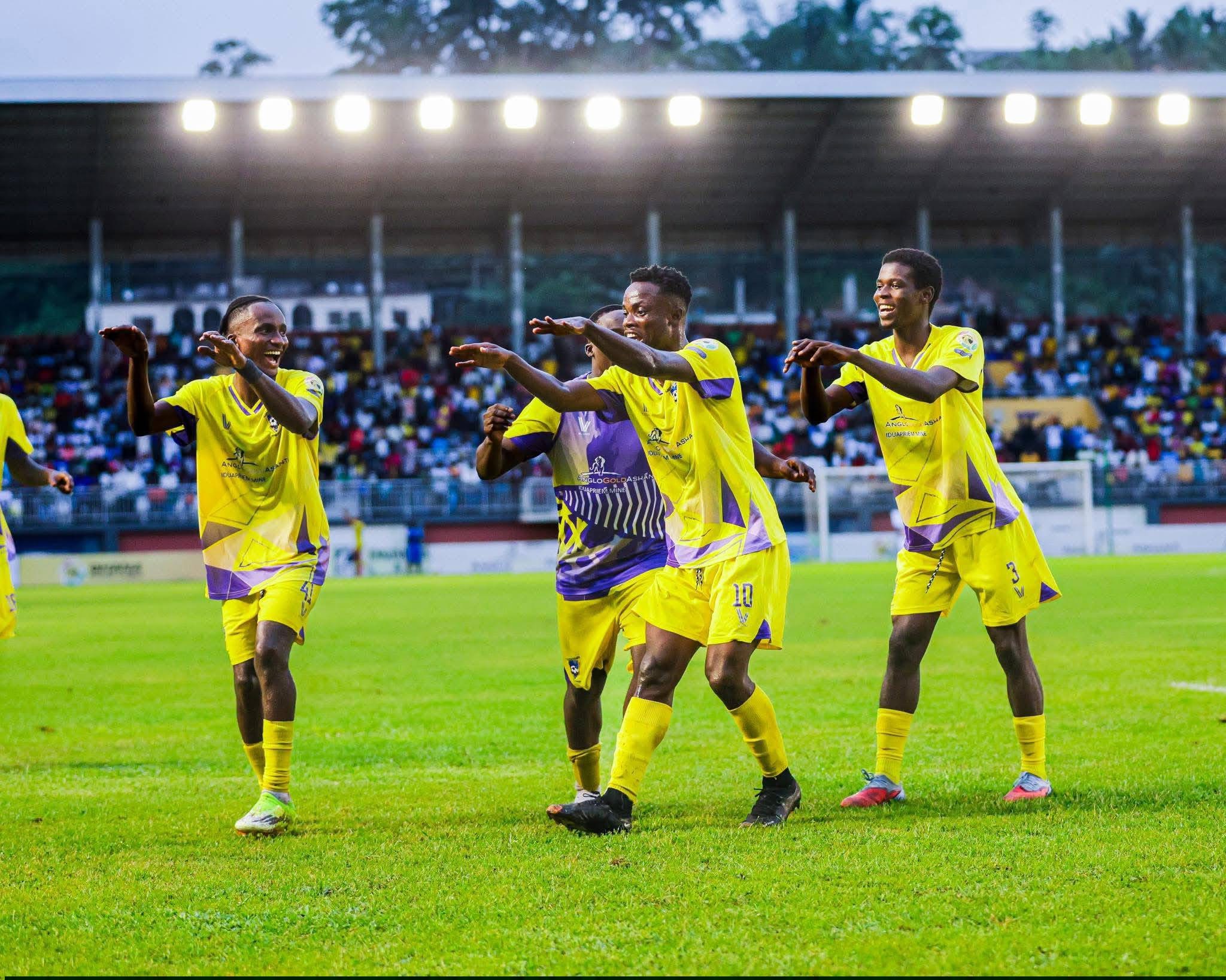Soccer players in bright yellow jerseys celebrate on the field with a cheering crowd in the stands behind them.