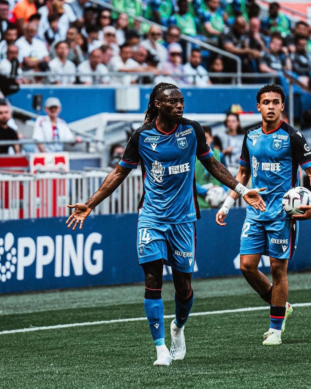 Two soccer players in blue uniforms on a green pitch, one with arms outstretched as a ball appears nearby, and a cheering crowd in the stands behind them for context of a match action scene near the sideline in a stadium.