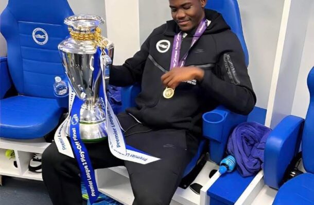 Athlete in a locker room wearing a medal, sitting in a blue chair, holding a large silver trophy with blue-and-white ribbons.