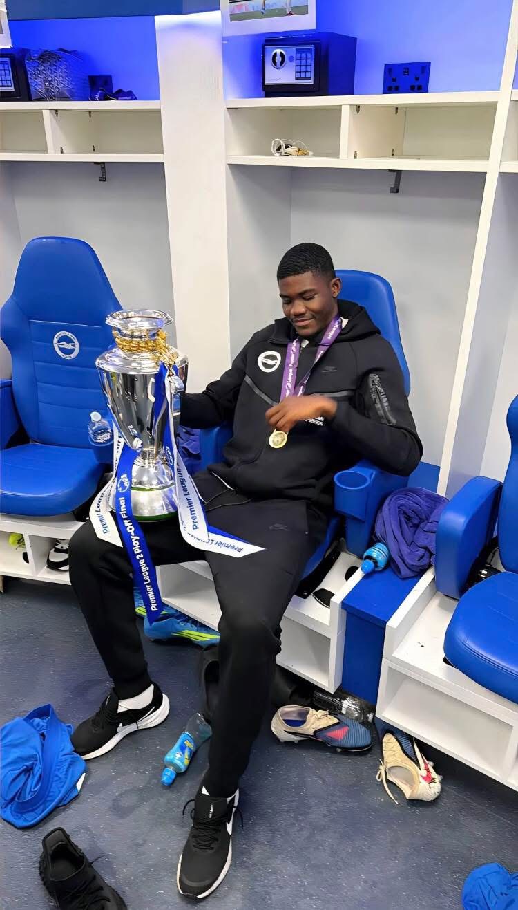 Athlete in a locker room wearing a medal, sitting in a blue chair, holding a large silver trophy with blue-and-white ribbons.
