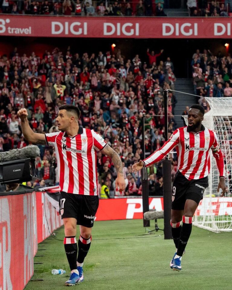 Two Atlético players in red-and-white striped jerseys celebrate near the sideline as fans cheer in the stands behind a goal.