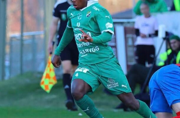 Soccer player in a green kit dribbles the ball along the field as a defender in blue lunges nearby during a match on a sunny day.