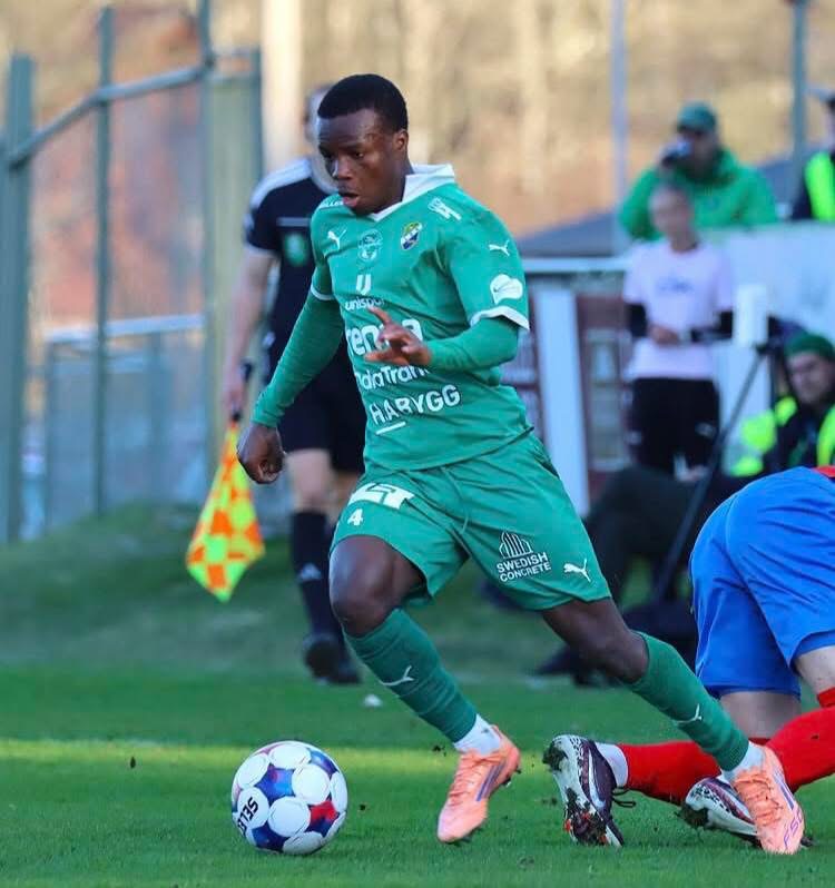 Soccer player in a green kit dribbles the ball along the field as a defender in blue lunges nearby during a match on a sunny day.