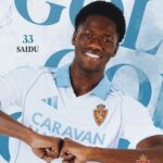 Smiling man in a light blue and white soccer jersey gesturing with fists, with ‘SAIDU 33’ in the background and team crest on sleeve.