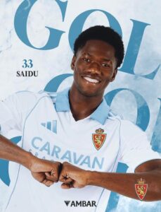 Smiling man in a light blue and white soccer jersey gesturing with fists, with ‘SAIDU 33’ in the background and team crest on sleeve.