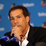 Man in a dark suit speaks at a press conference, hand resting near his mouth with multiple microphones in front of him.