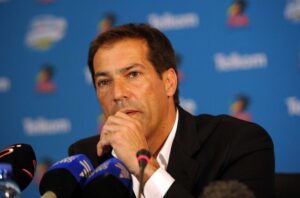 Man in a dark suit speaks at a press conference, hand resting near his mouth with multiple microphones in front of him.