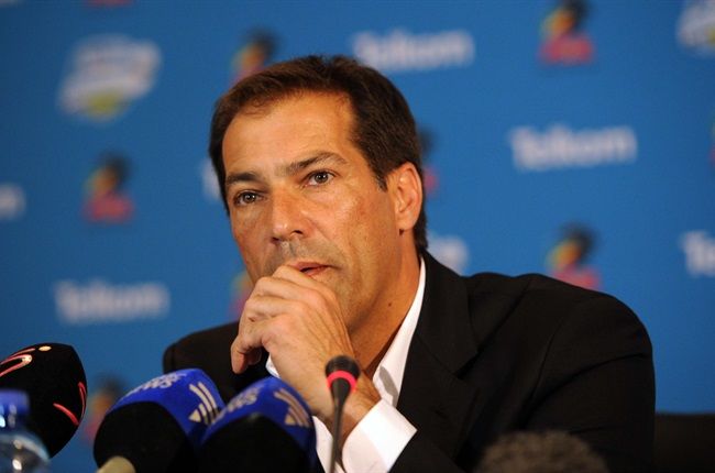 Man in a dark suit speaks at a press conference, hand resting near his mouth with multiple microphones in front of him.
