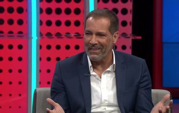 Smiling man in a navy suit and white shirt sits on a gray chair, gesturing with his hands during a television interview set with pink dotted background and blue lighting.