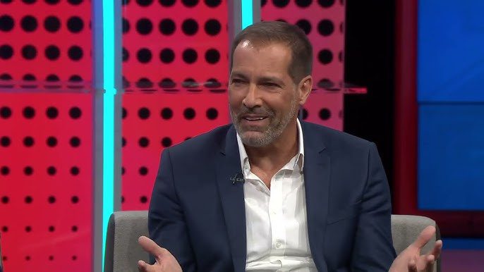 Smiling man in a navy suit and white shirt sits on a gray chair, gesturing with his hands during a television interview set with pink dotted background and blue lighting.