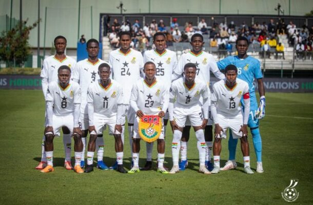 Eleven soccer players in white uniforms pose for a group photo on a green field, with a blue-clad goalkeeper on the right and fans in the stands behind them.
