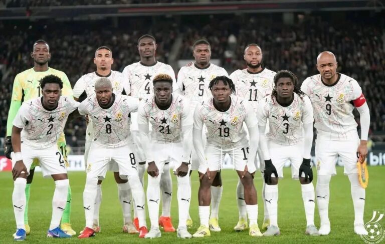 Eleven soccer players in white uniforms pose for a team lineup on the field, with a neon-green-clad goalkeeper on the left and a crowd in the stands behind them.