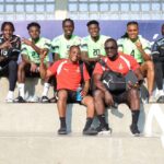 Group of soccer players and two staff members sit on stadium steps, posing for a photo in green jerseys and red shirts.