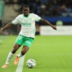 Soccer player in white and green kit controls the ball on a grass field during a match, number 20 visible on shorts