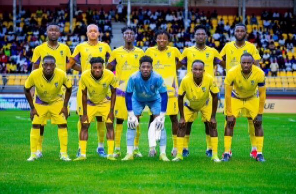 IMG_0609 | Ghanaguardian.com Soccer team in yellow uniforms posing for a group photo on the field with spectators in the stands behind them, goalkeeper in blue front center.