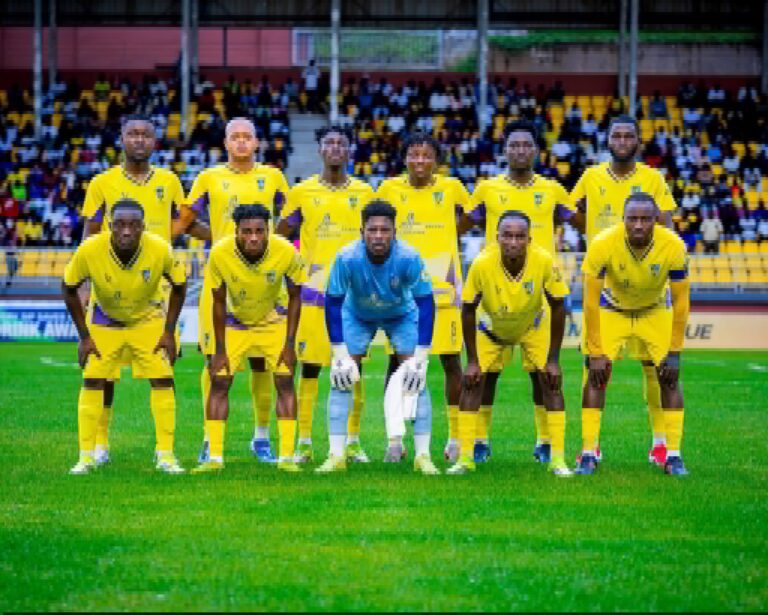 Soccer team in yellow uniforms posing for a group photo on the field with spectators in the stands behind them, goalkeeper in blue front center.