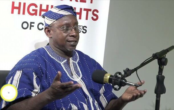 Man in blue traditional clothing speaking into a microphone at a press event, banner behind him reads 'HIGHLIGHTS'.