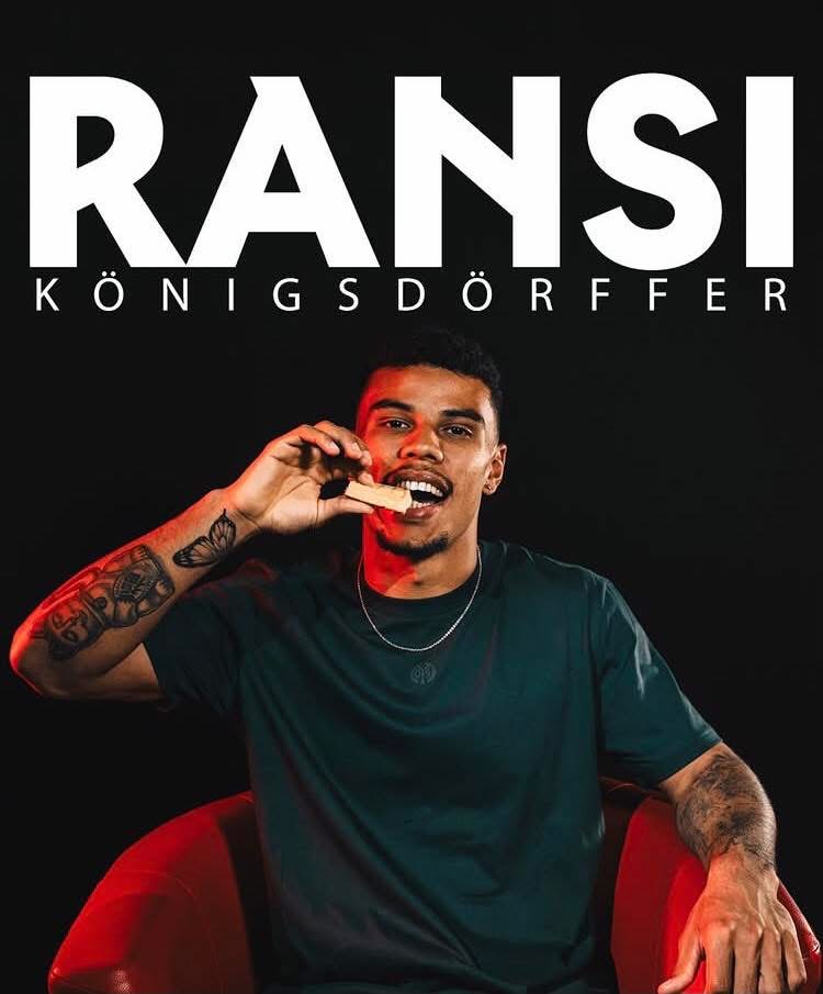 Portrait of a man with tattoos biting a snack while seated in a red chair, dark background and large 'RANSI' text above.
