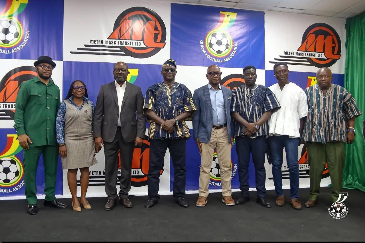 Nine people posing for a group photo at a football association event, with banners displaying soccer logos behind them.