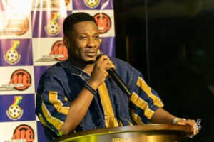 Man in blue and yellow traditional attire speaks into a handheld microphone at a podium before a backdrop of logos on a patterned wall.