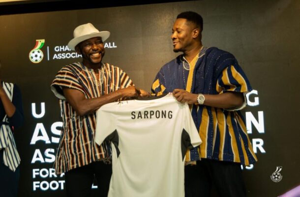 Two men on a stage shake hands while presenting a white soccer jersey that reads ‘SARPONG,’ smiling at the moment of the handoff.