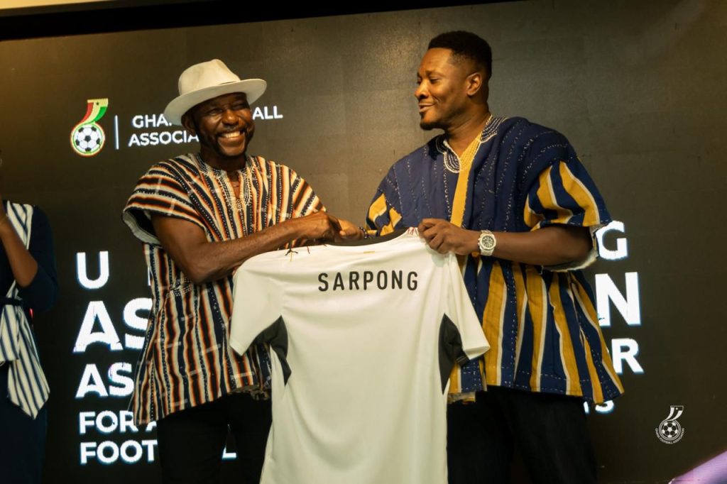 Two men on a stage shake hands while presenting a white soccer jersey that reads ‘SARPONG,’ smiling at the moment of the handoff.