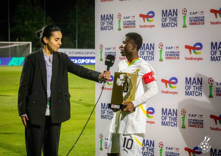Reporter interviews a soccer player in a white jersey with a trophy on a podium backdrop that reads 'Man of the Match'.