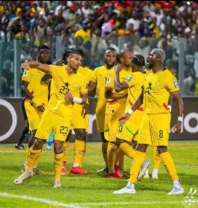 Soccer players in yellow uniforms celebrate on the field, group huddle with fans in the stadium behind.