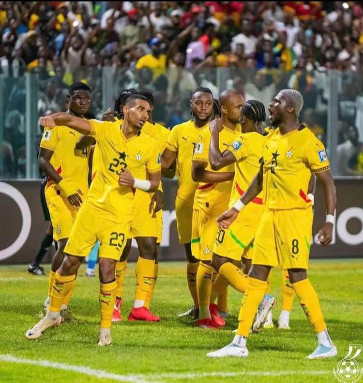 Soccer players in yellow uniforms celebrate on the field, group huddle with fans in the stadium behind.