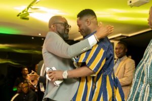 Two men hug warmly at a lively indoor event, one in a light gray suit and glasses, the other in a blue-and-gold striped outfit, while guests watch in the background.