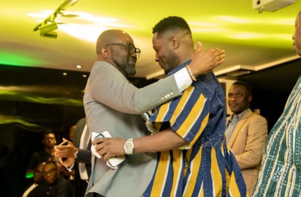 Two men hug warmly at a lively indoor event, one in a light gray suit and glasses, the other in a blue-and-gold striped outfit, while guests watch in the background.