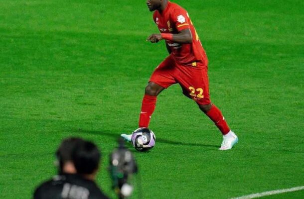 Soccer player in a red kit (number 22) dribbles the ball on a green pitch, with a photographer in the foreground recording the action.