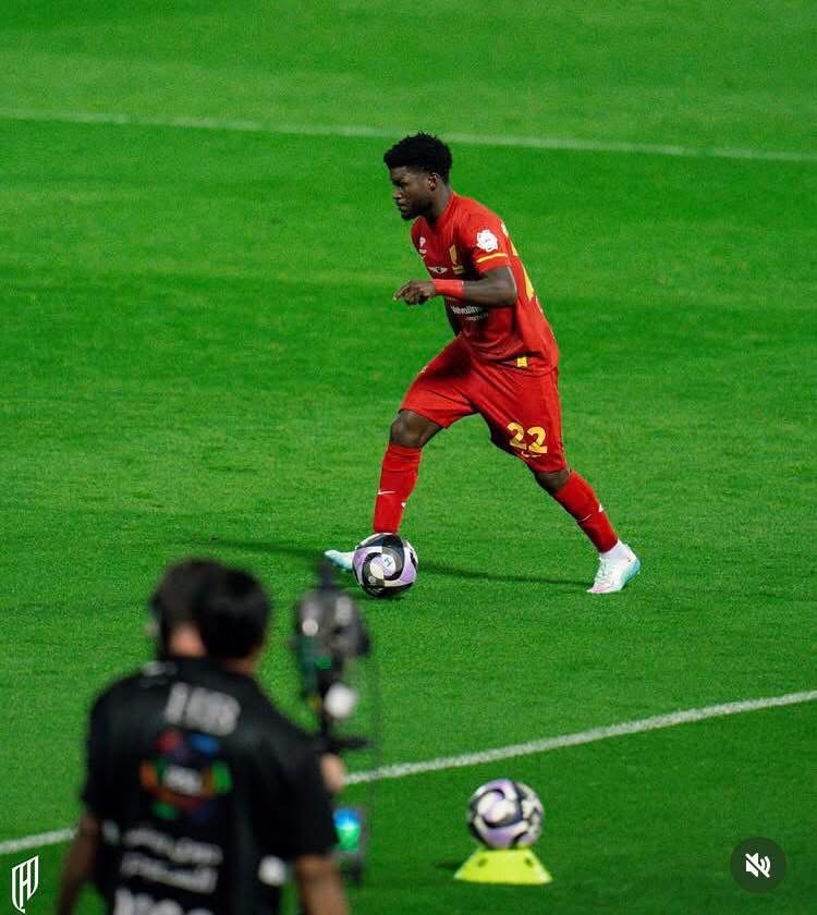 Soccer player in a red kit (number 22) dribbles the ball on a green pitch, with a photographer in the foreground recording the action.