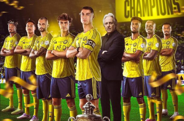 Soccer team in yellow uniforms posing on a lit stadium field with a large 'CHAMPIONS' banner in back; a coach stands among players and a trophy sits at the foreground.