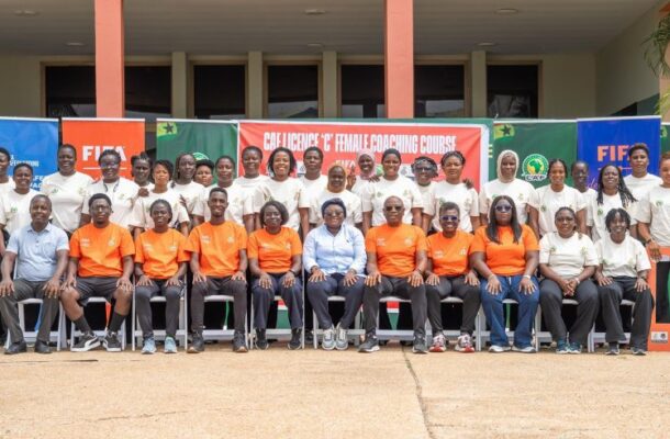 Group of men and women in orange and white shirts posing for a photo in front of a FIFA coaching course banner outdoors.